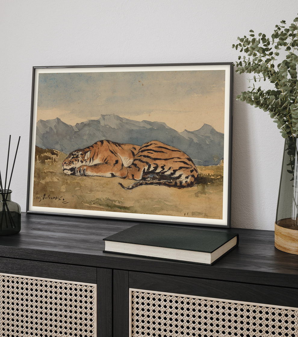 Framed watercolor painting by Eugène Delacroix depicting a resting tiger in a landscape with distant mountains, displayed on a dark wood cabinet with rattan panels.