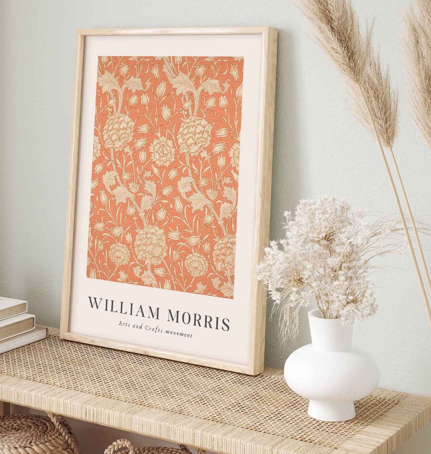 Framed William Morris art print with an orange and beige floral pattern from the Arts and Crafts movement, displayed on a woven console beside a white vase of dried flowers.