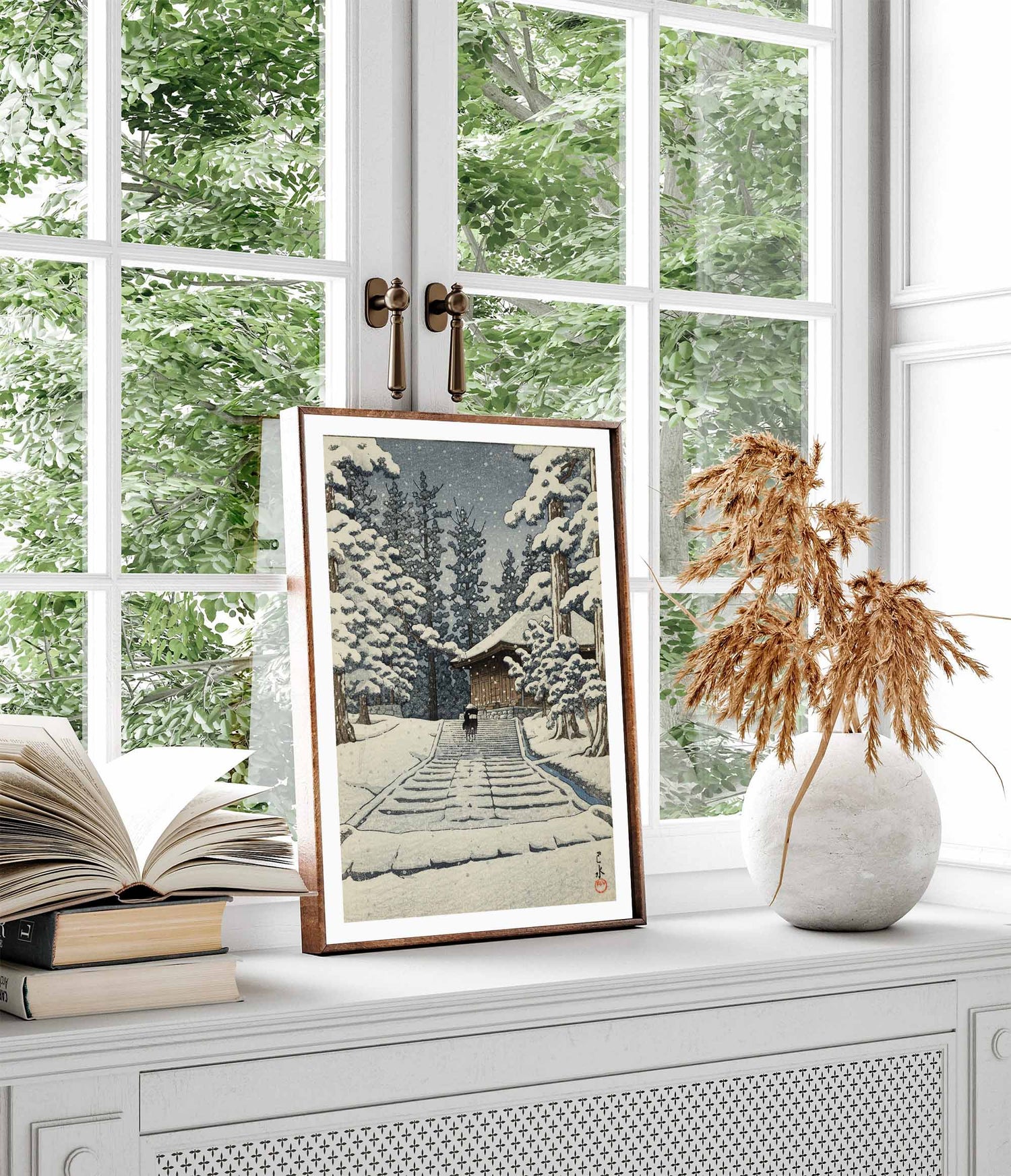 Framed art print of Kawase Hasui’s “Hiraizumi, Konjikidō in Snow,” showing a snowy temple path lined with trees, displayed on a windowsill beside books and dried flowers, with greenery visible through the window.