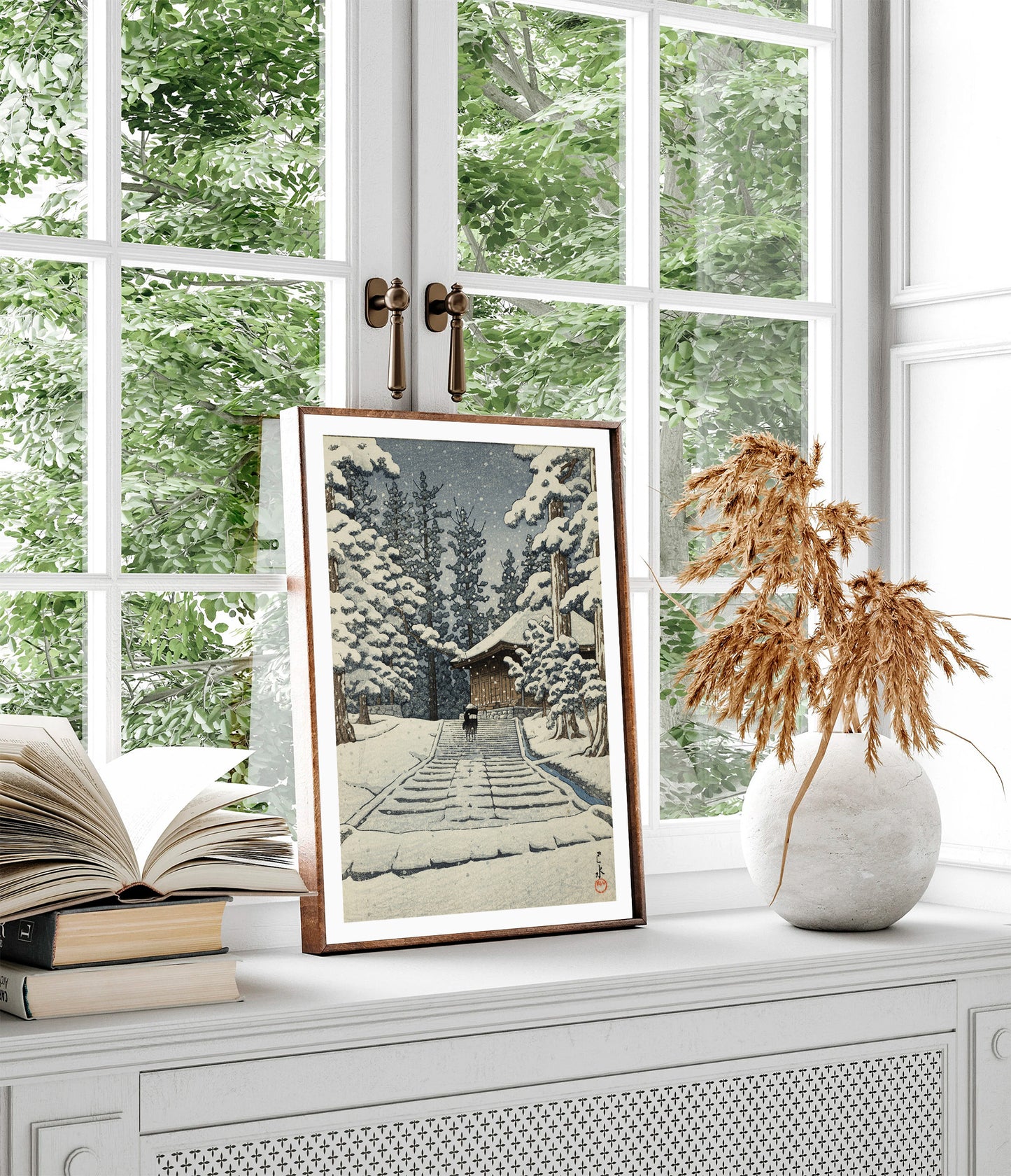Framed art print of Kawase Hasui’s “Hiraizumi, Konjikidō in Snow,” showing a snowy temple path lined with trees, displayed on a windowsill beside books and dried flowers, with greenery visible through the window.