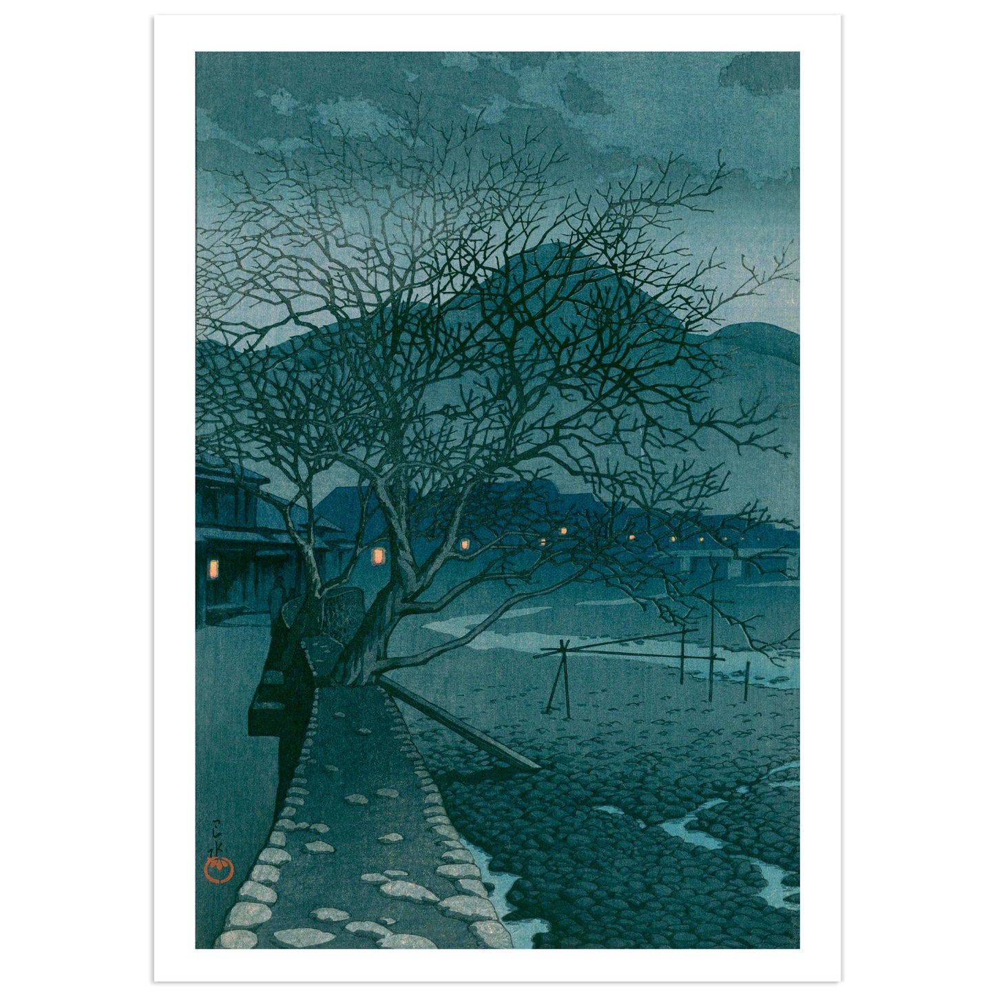 Japanese shin-hanga woodblock print by Kawase Hasui showing a stone path curving along a boulder-strewn riverbed at dusk in the hot-spring town of Beppu, with two bare winter trees overhead, blue hills behind, and small orange lantern lights glowing in the inns along the far bank.