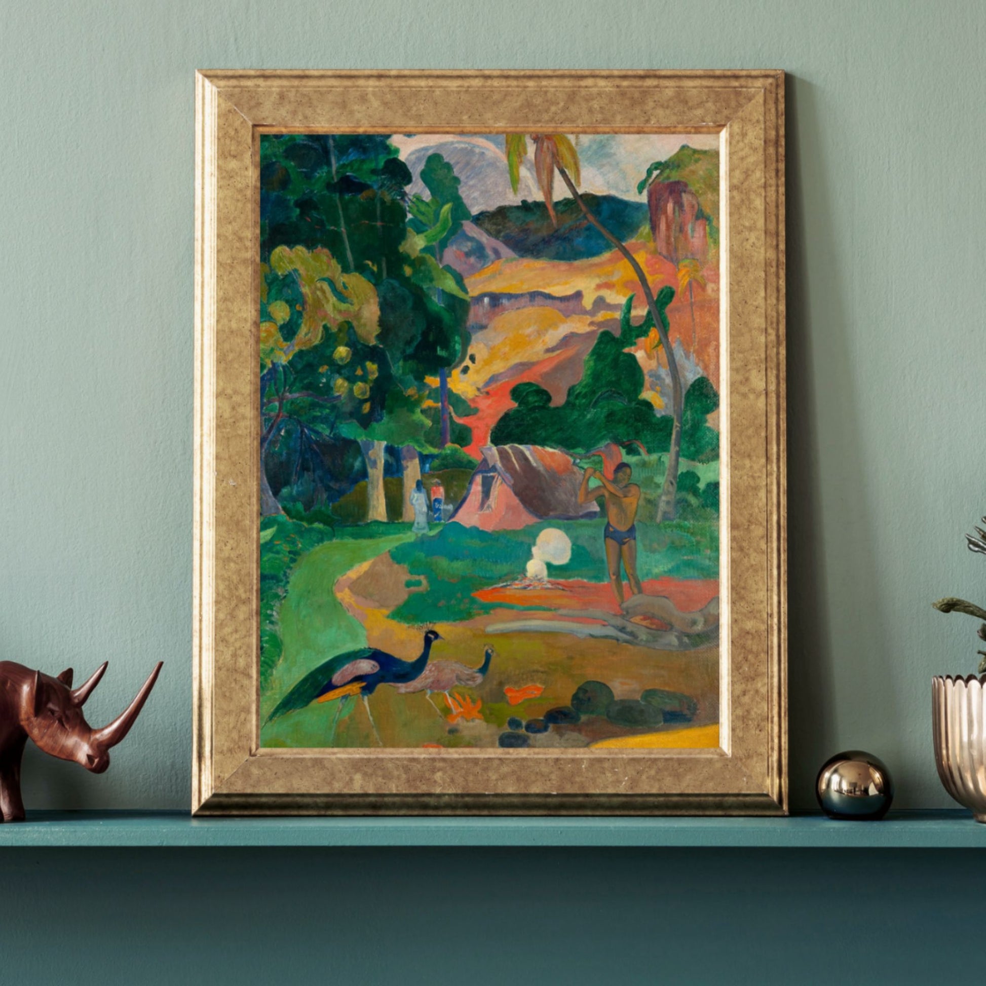 A framed poster of a Paul Gauguin painting titled 'Matamoe (Death), Landscape with Peacocks', displayed on a blue wall above a mantelpiece with a decorative rhinoceros and a plant.