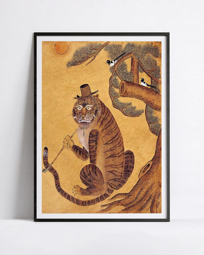 A framed art print featuring a whimsical illustration of a minhwa tiger wearing a top hat and holding a cane, seated on a tree branch against a warm gold background. The print is displayed in a black frame with a white mat, leaning against a light gray wall.