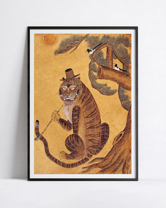 A framed art print featuring a whimsical illustration of a minhwa tiger wearing a top hat and holding a cane, seated on a tree branch against a warm gold background. The print is displayed in a black frame with a white mat, leaning against a light gray wall.