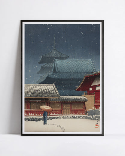 A framed art print featuring a Kawase Hasui shin-hanga print snow scene with traditional temple buildings, a red gate, and a person holding an umbrella in the foreground, displayed against a light gray wall.
