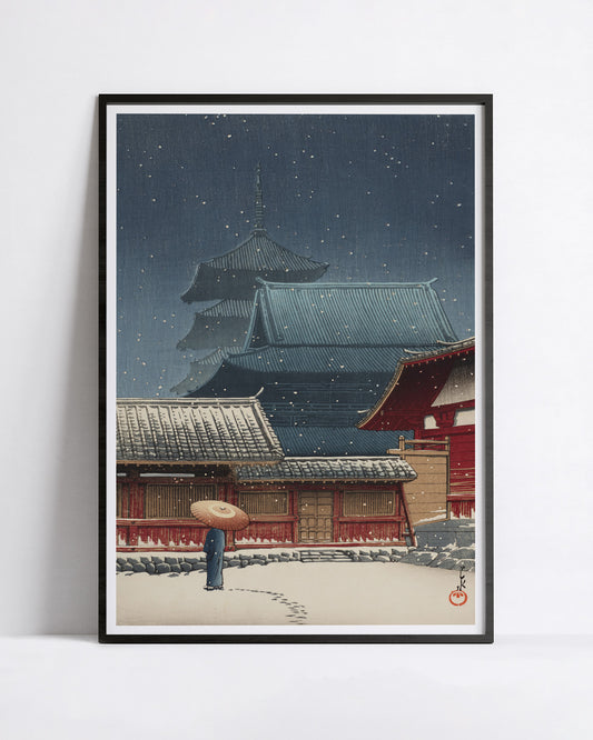 A framed art print featuring a Kawase Hasui shin-hanga print snow scene with traditional temple buildings, a red gate, and a person holding an umbrella in the foreground, displayed against a light gray wall.